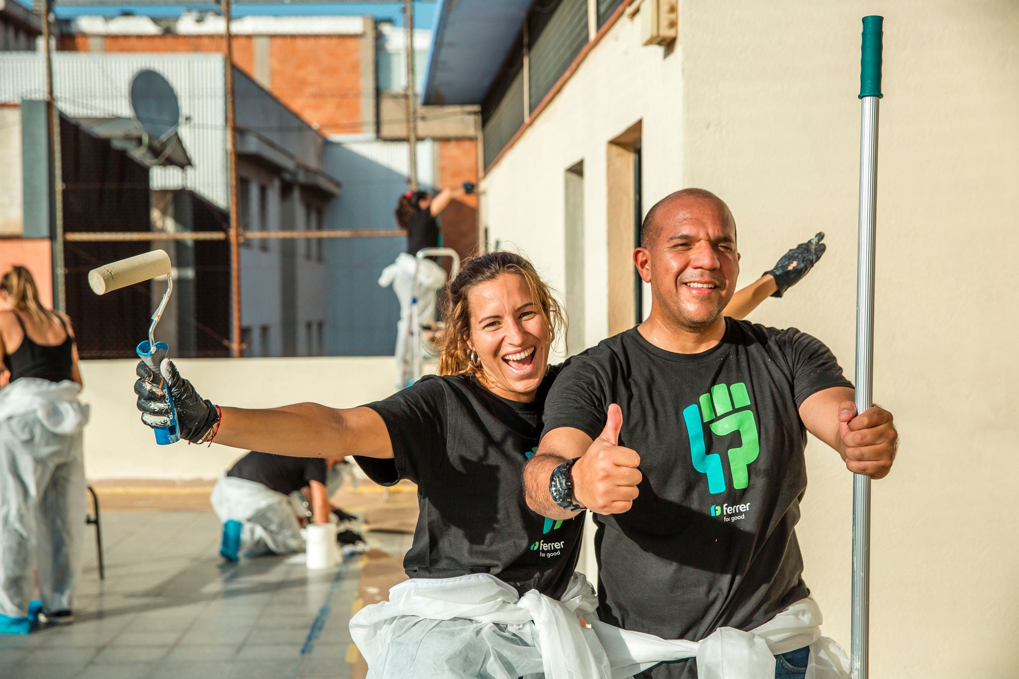 Two smiling volunteers in corporate T-shirts celebrate as they participate in a community painting activity on a rooftop.