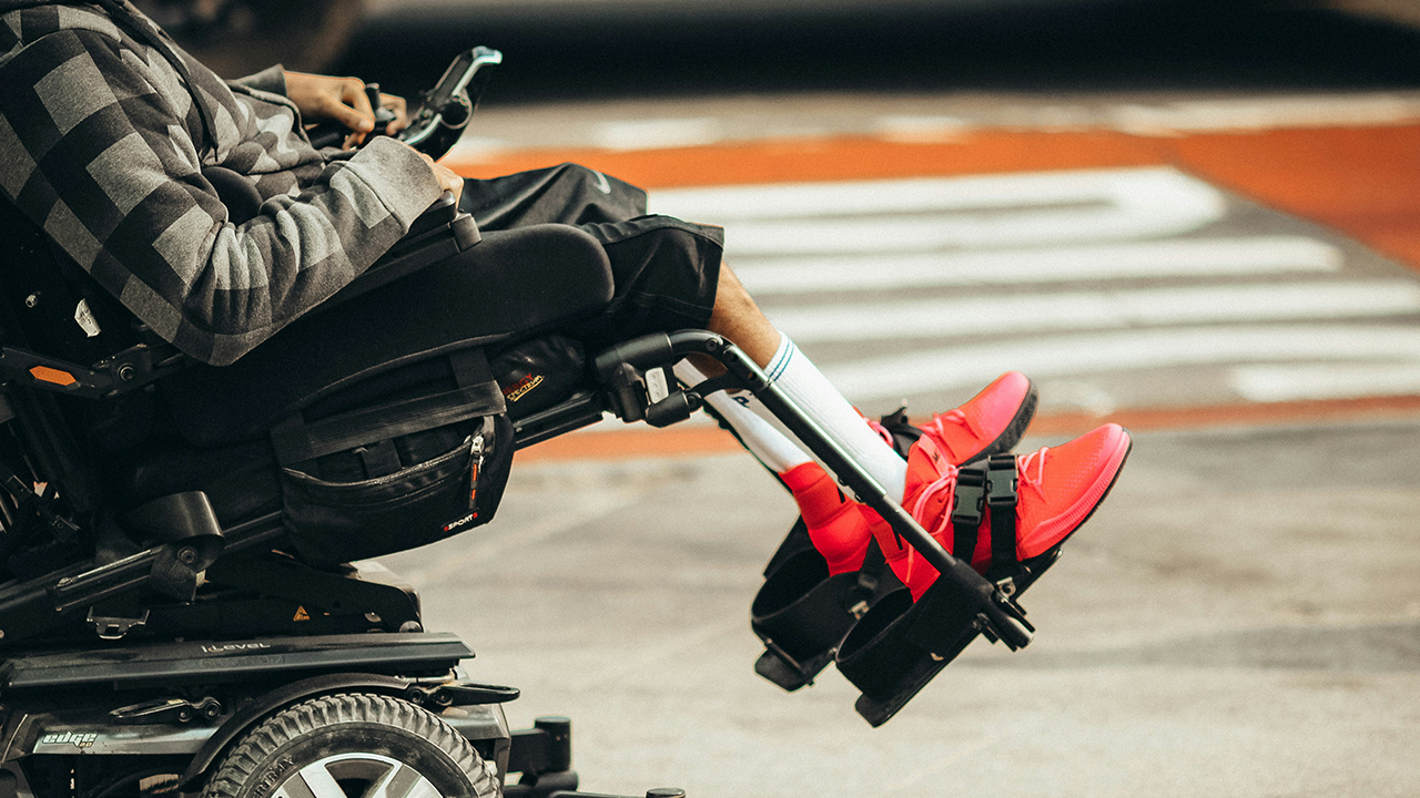 Profile view and from a low angle of a person in bright red sneakers sitting in a motorized wheelchair on the street.