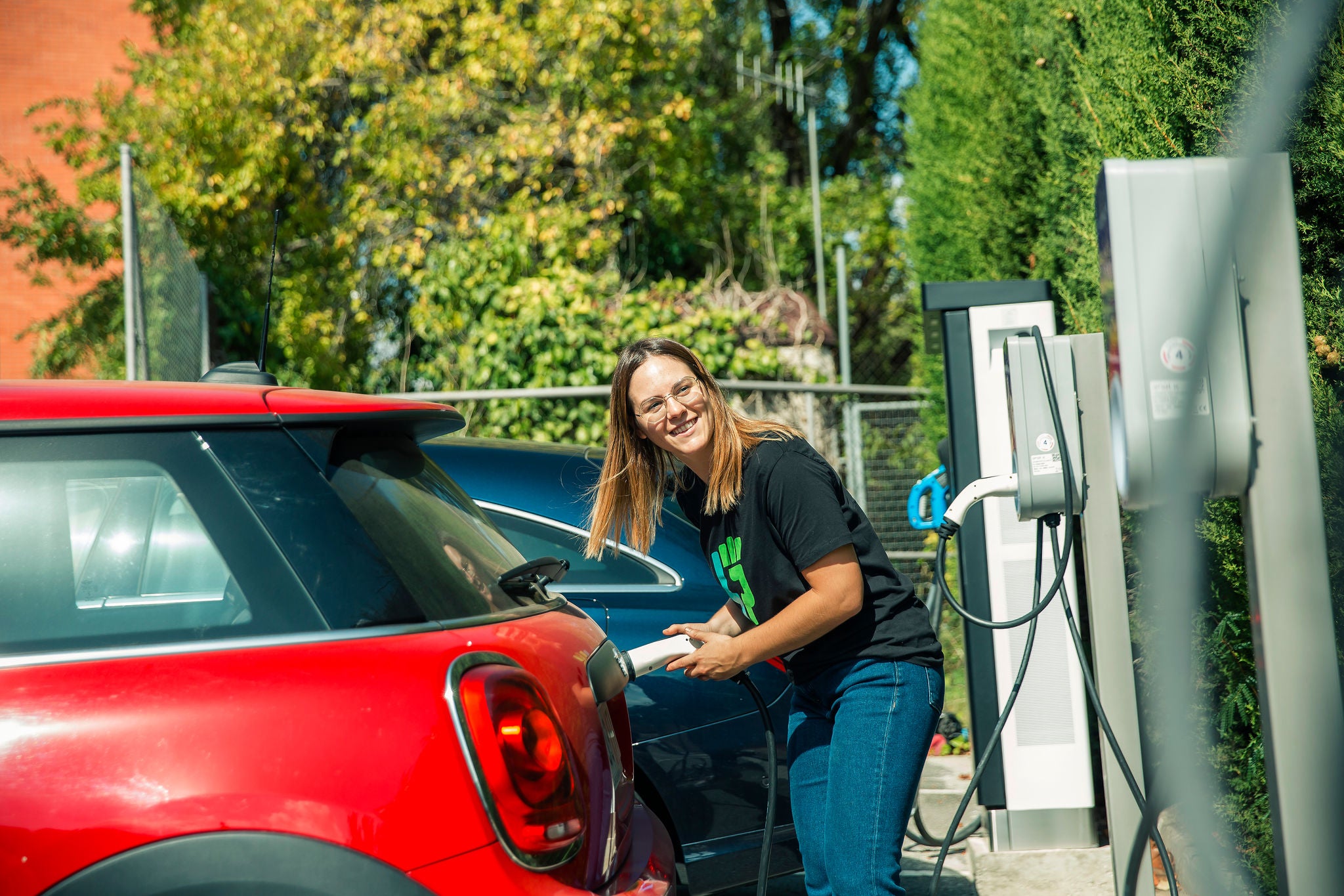 A woman smiles as she connects the cable from a charging station to a red electric car parked outdoors.
