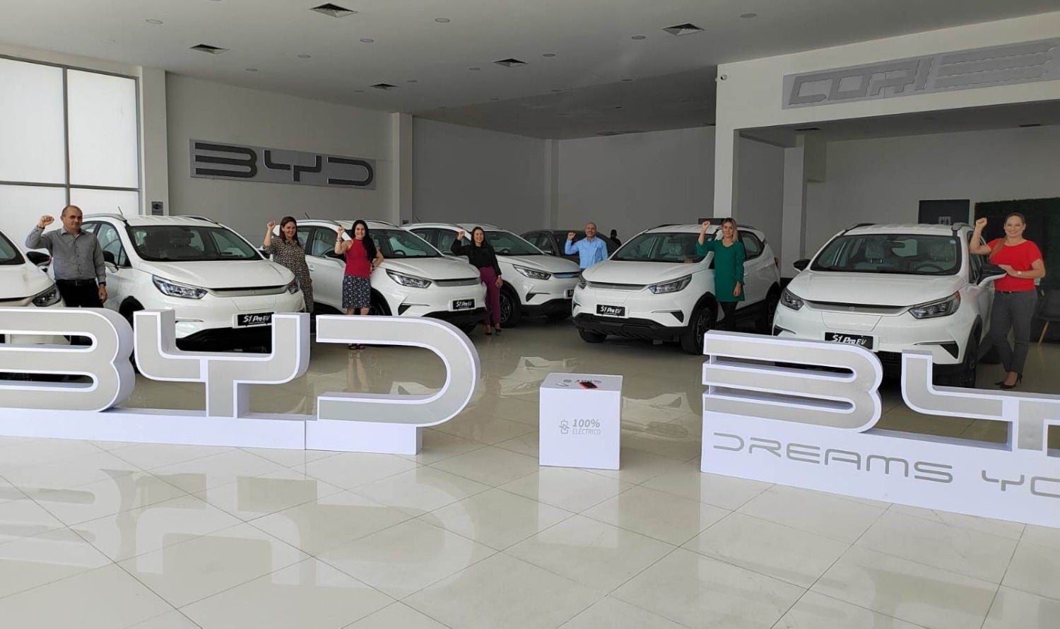 Concessionari de cotxes BYD amb diversos vehicles elèctrics blancs exposats i un grup de persones posant al costat d'ells amb el puny aixecat.