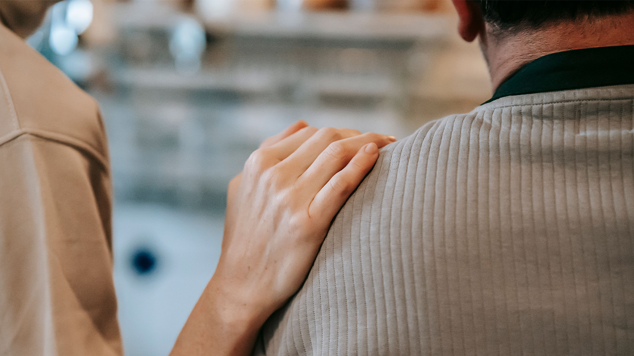 Rear view of a person gently placing a hand on the shoulder of another dressed in a gray sweater, in a gesture of comfort or support.