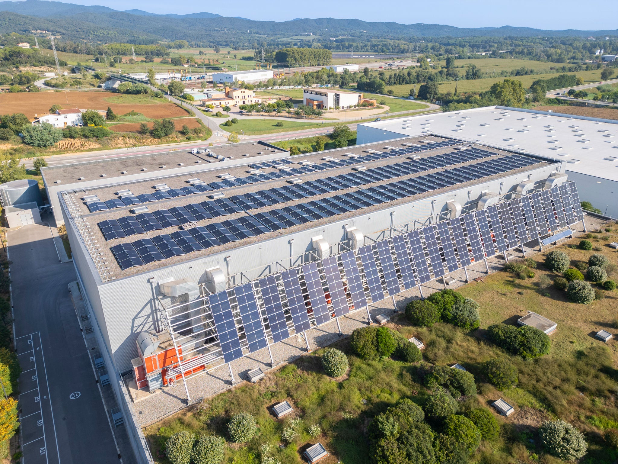 Vista aèria d'una nau industrial amb una gran instal·lació de panells solars fotovoltaics a la teulada i a la façana lateral.