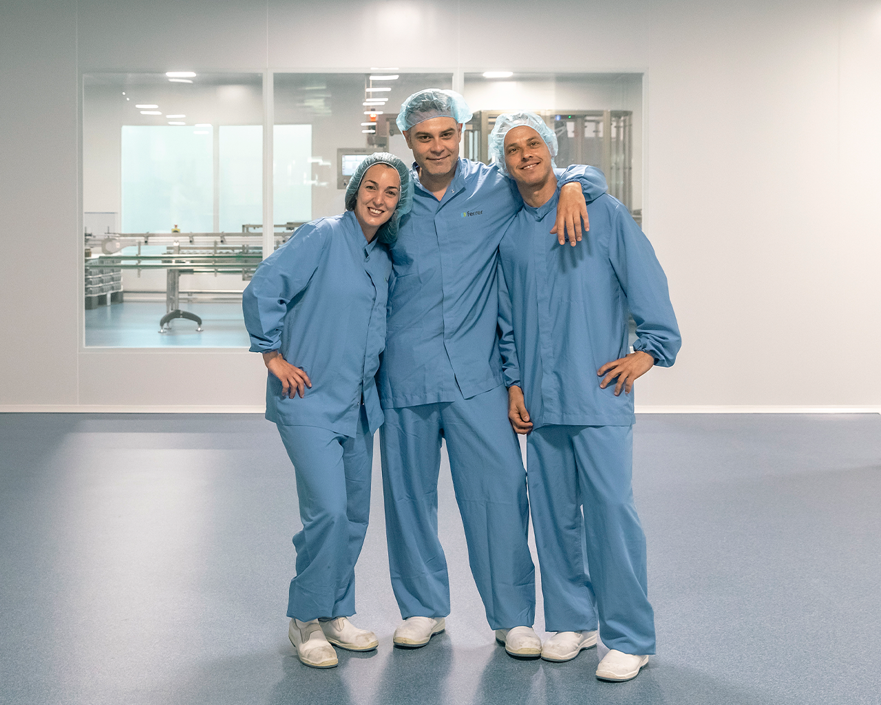 Equipo de tres operarios con trajes de protección, reflejando el trabajo en equipo, la cultura y los valores de Grupo Ferrer en su planta de producción farmacéutica sostenible.
