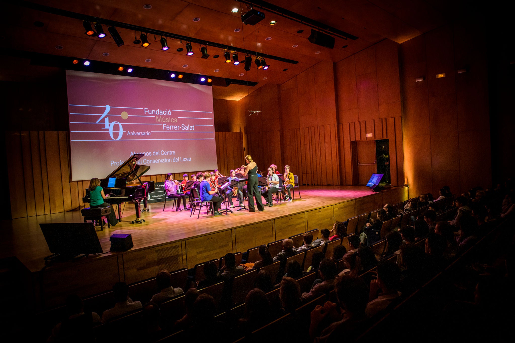 Joves músics assagen en un auditori de fusta davant el públic, sota una pantalla que celebra el 40è aniversari de la Fundació Música Ferrer-Salat.