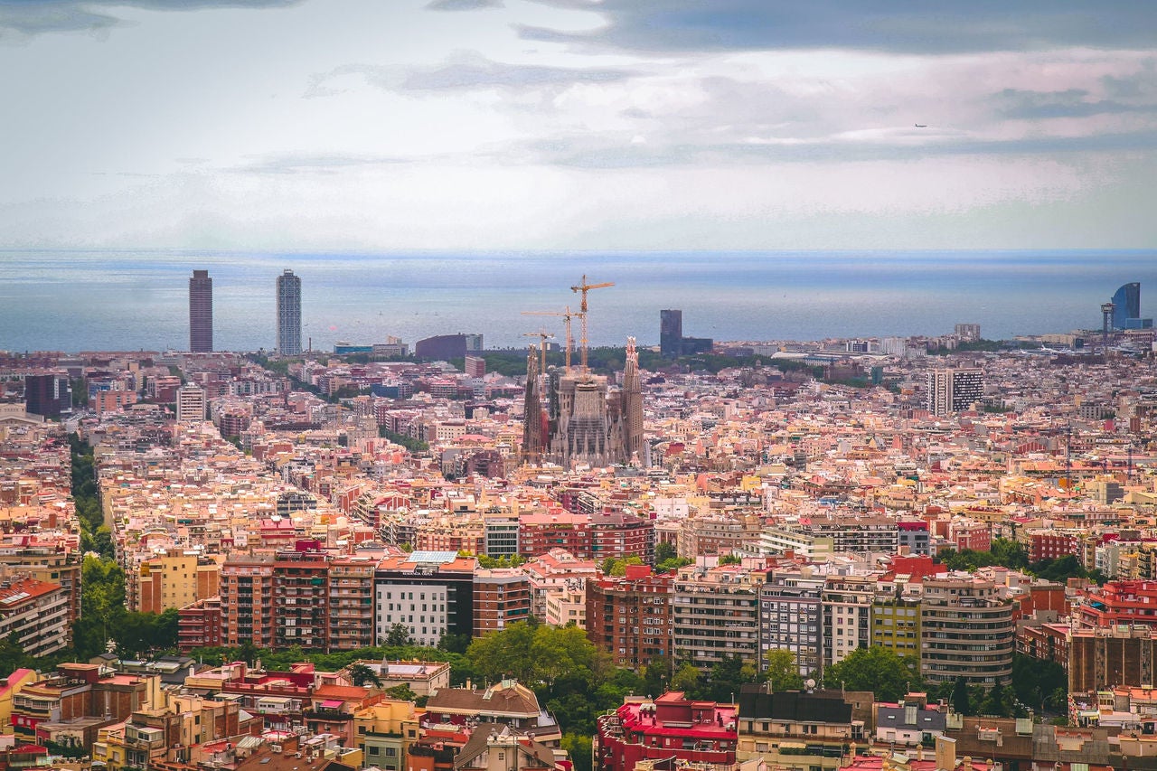 Vista panorámica de Barcelona con la Sagrada Familia en el centro, el mar al fondo y el perfil urbano extendiéndose bajo un cielo nublado.