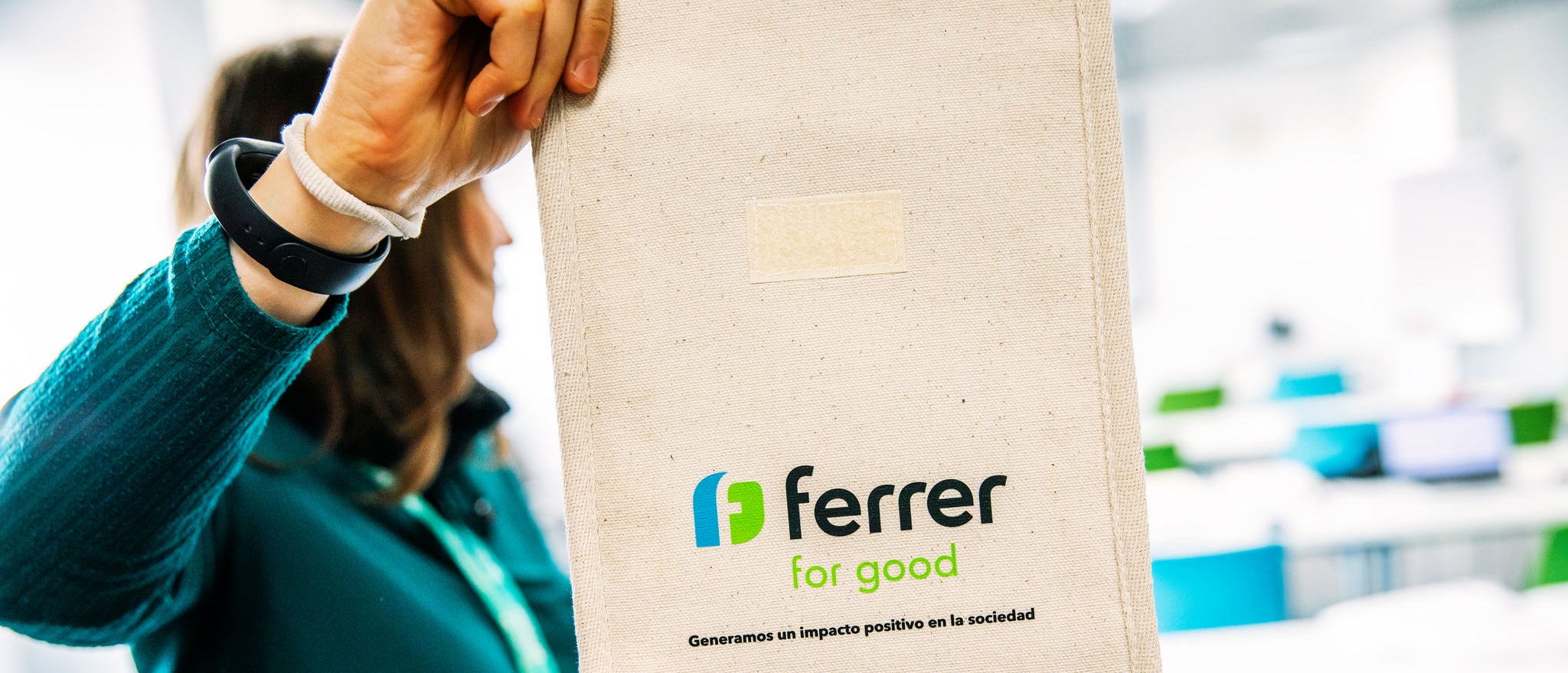 Person holds a reusable cloth bag with the "Ferrer for Good" logo in a bright interior space, related to a Ferrer corporate or volunteer activity.