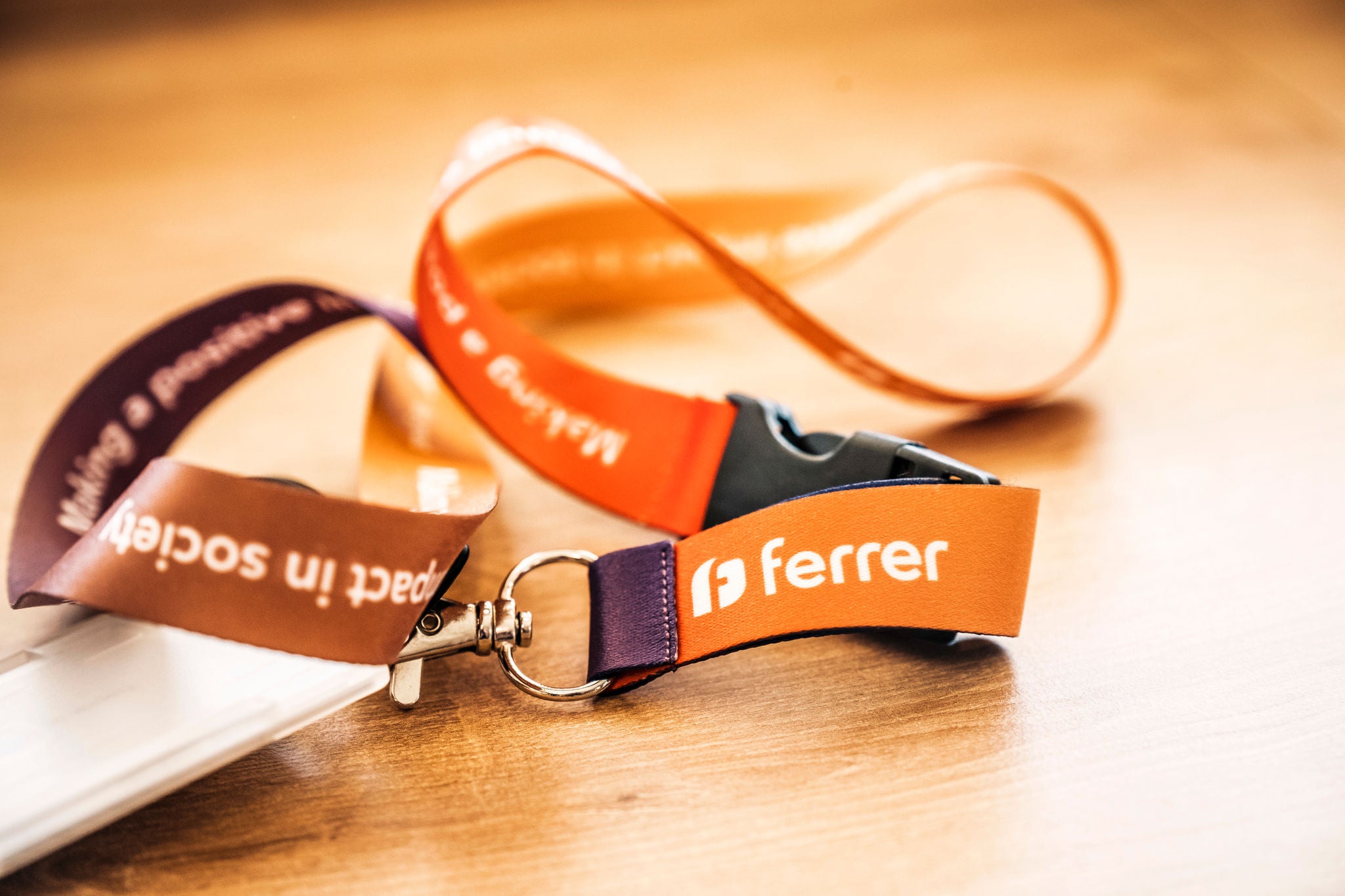 Cordón identificativo naranja sobre una mesa, con el logotipo “ferrer” impreso en blanco y un clip metálico en primer plano.