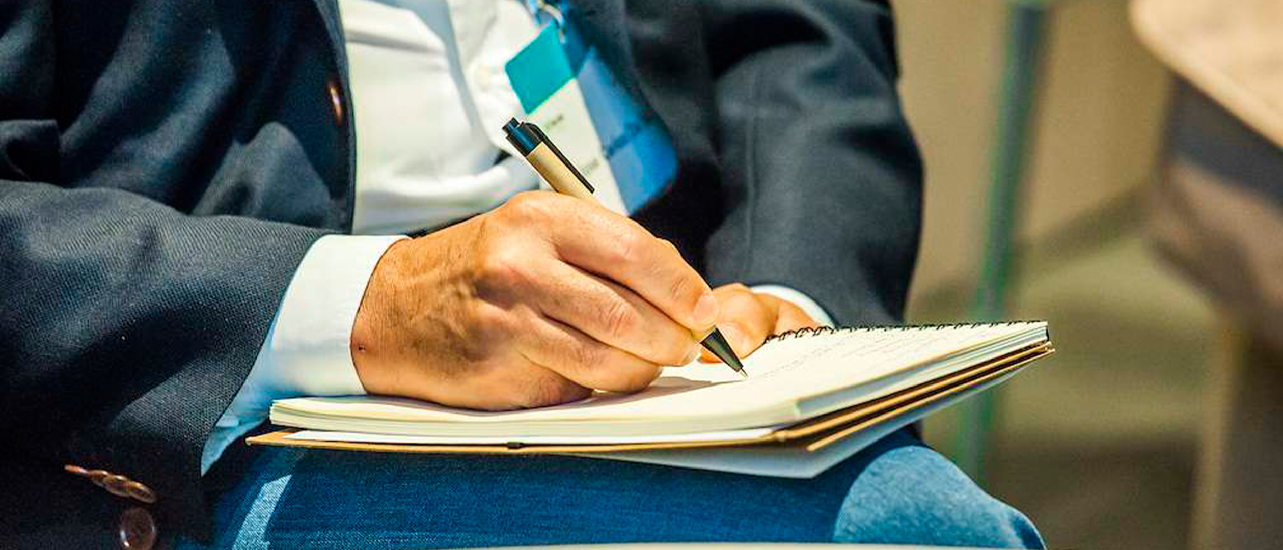 Primer pla d'una persona amb jaqueta fosca prenent notes amb un bolígraf en una llibreta recolzada sobre les seves cames durant un esdeveniment corporatiu o conferència.