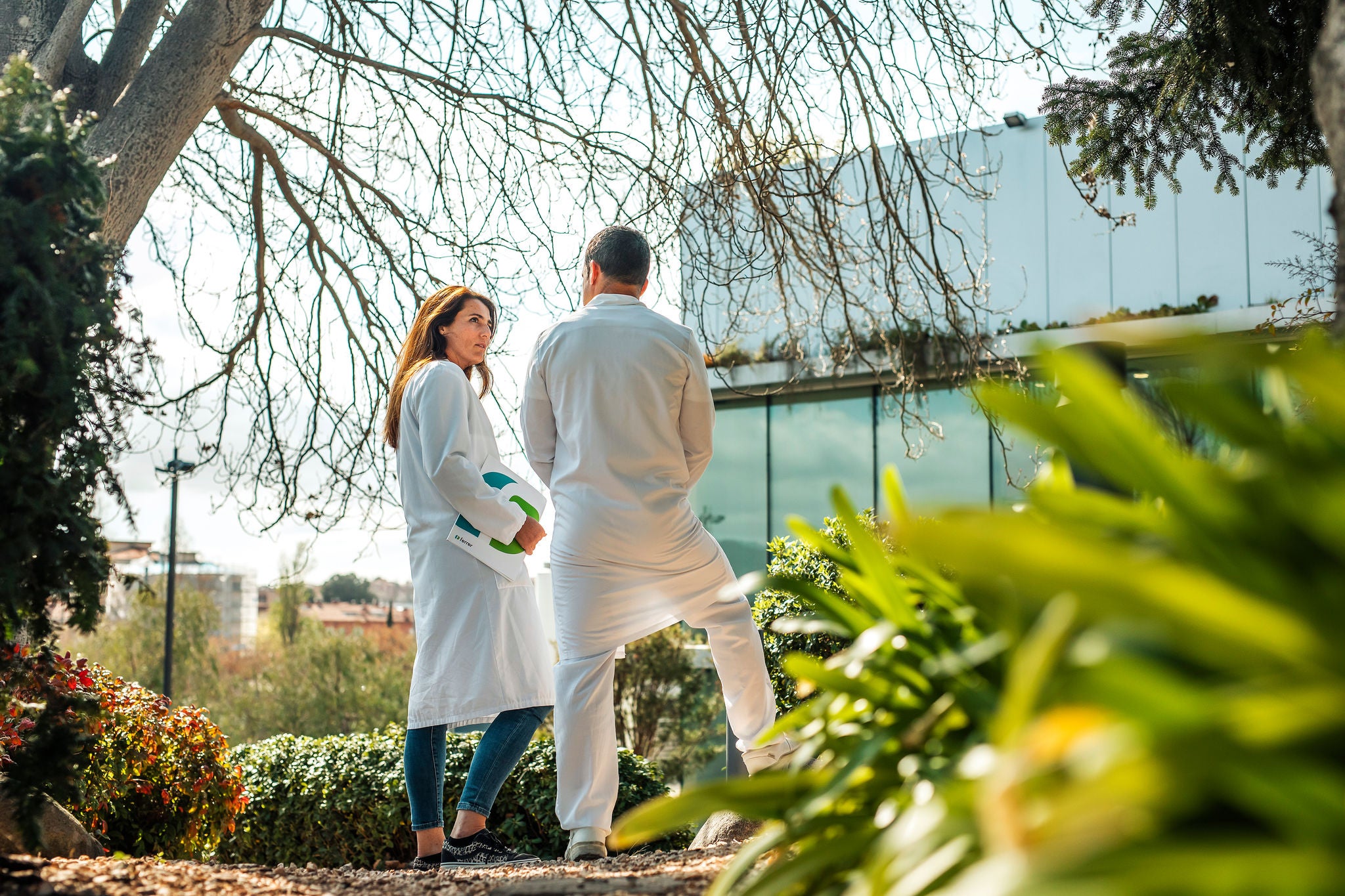 Dos professionals sanitaris amb bata blanca conversen en un jardí a l'aire lliure davant un edifici modern.