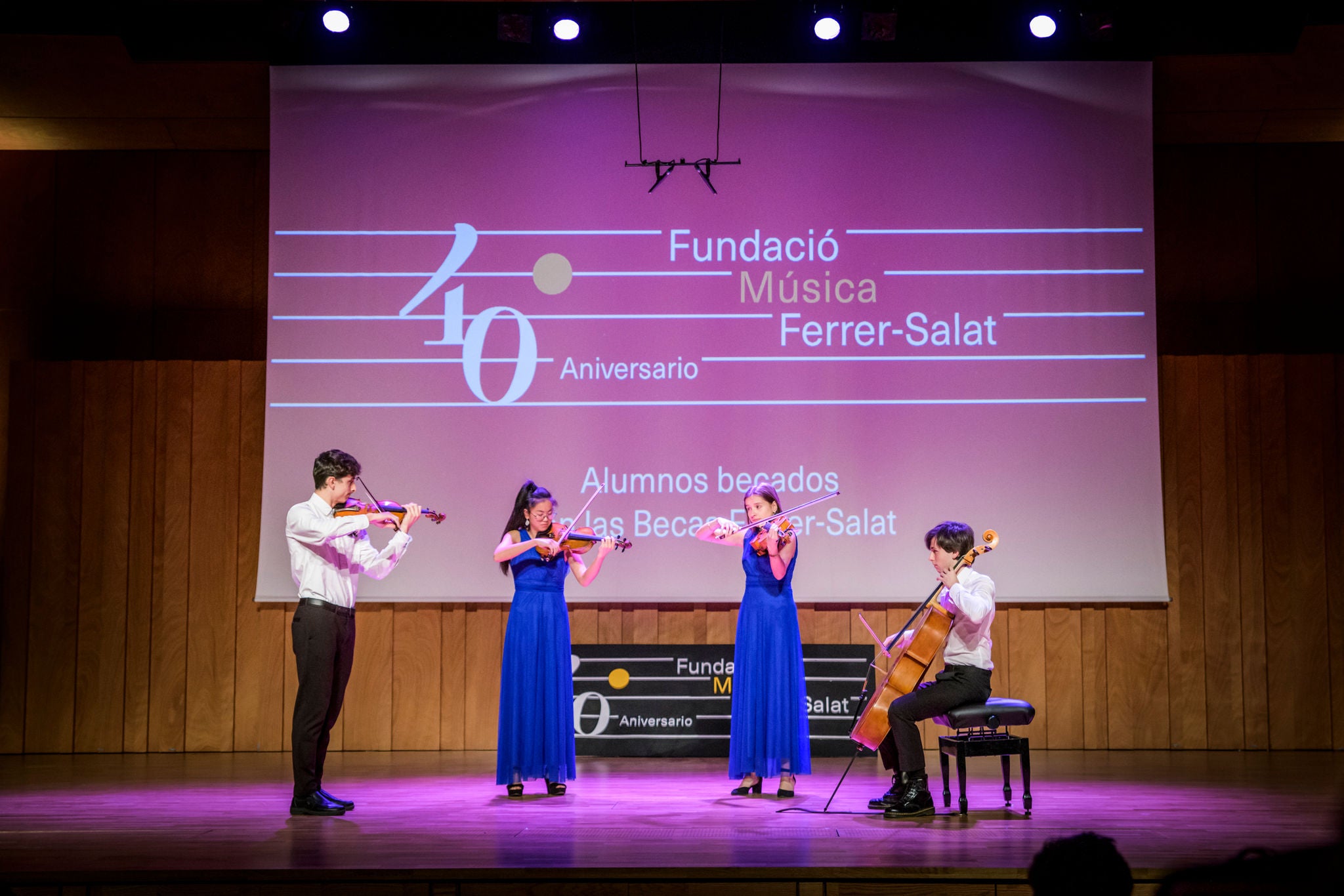String quartet composed of three standing musicians and a cellist seated performing on a stage in front of a commemorative screen of the Fundació Música Ferrer-Salat.
