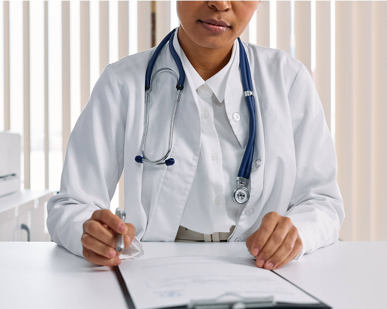 A doctor in a white coat and stethoscope holds a pen over a medical history at her desk.