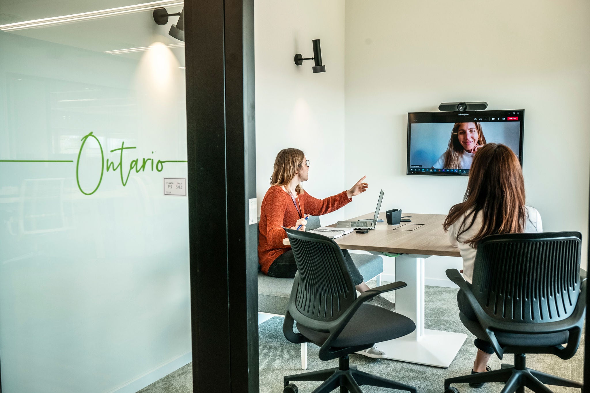 Two people hold a video conference from a modern meeting room called "Ontario."