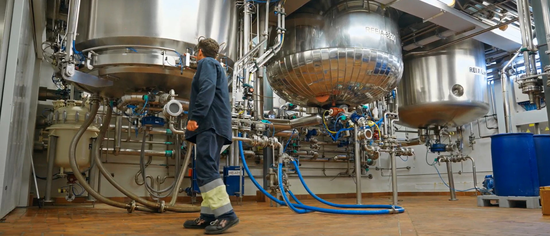 Professional de Ferrer en una zona de producció industrial, caminant al costat d'equips i dipòsits metàl·lics.