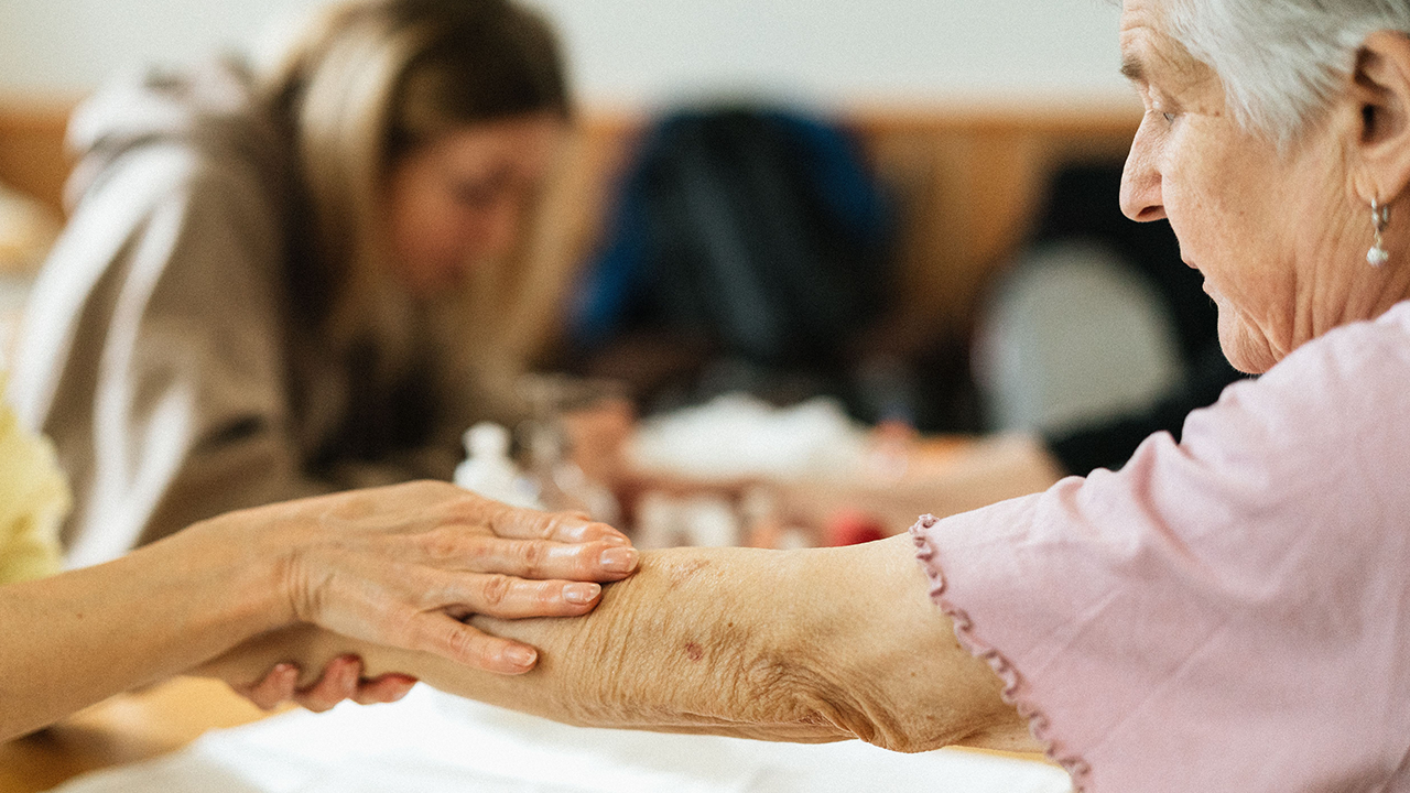 A young hand gently holds the arm of an older woman with gray hair.