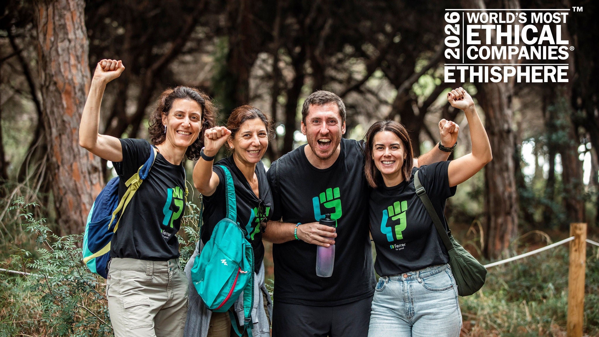 Four smiling people in matching T-shirts celebrating in a forest with their fists raised.