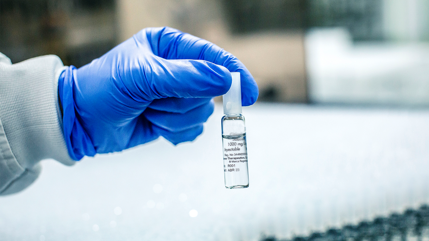 Primer pla d' una mà amb guant blau sostenint una ampolla de medicament injectable a la planta de producció de Ferrer.