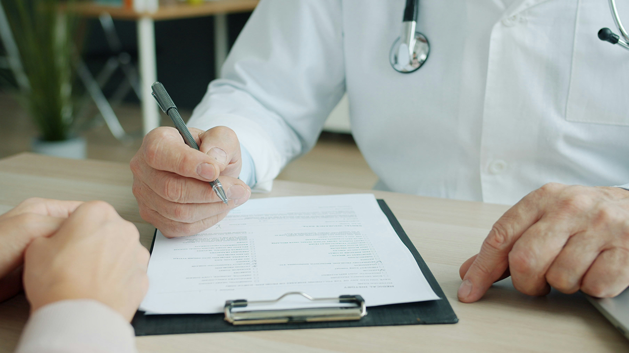 Primer plano de un médico en bata blanca con un estetoscopio sosteniendo un bolígrafo sobre un historial clínico en una tabla portapapeles mientras atiende a un paciente en su escritorio.