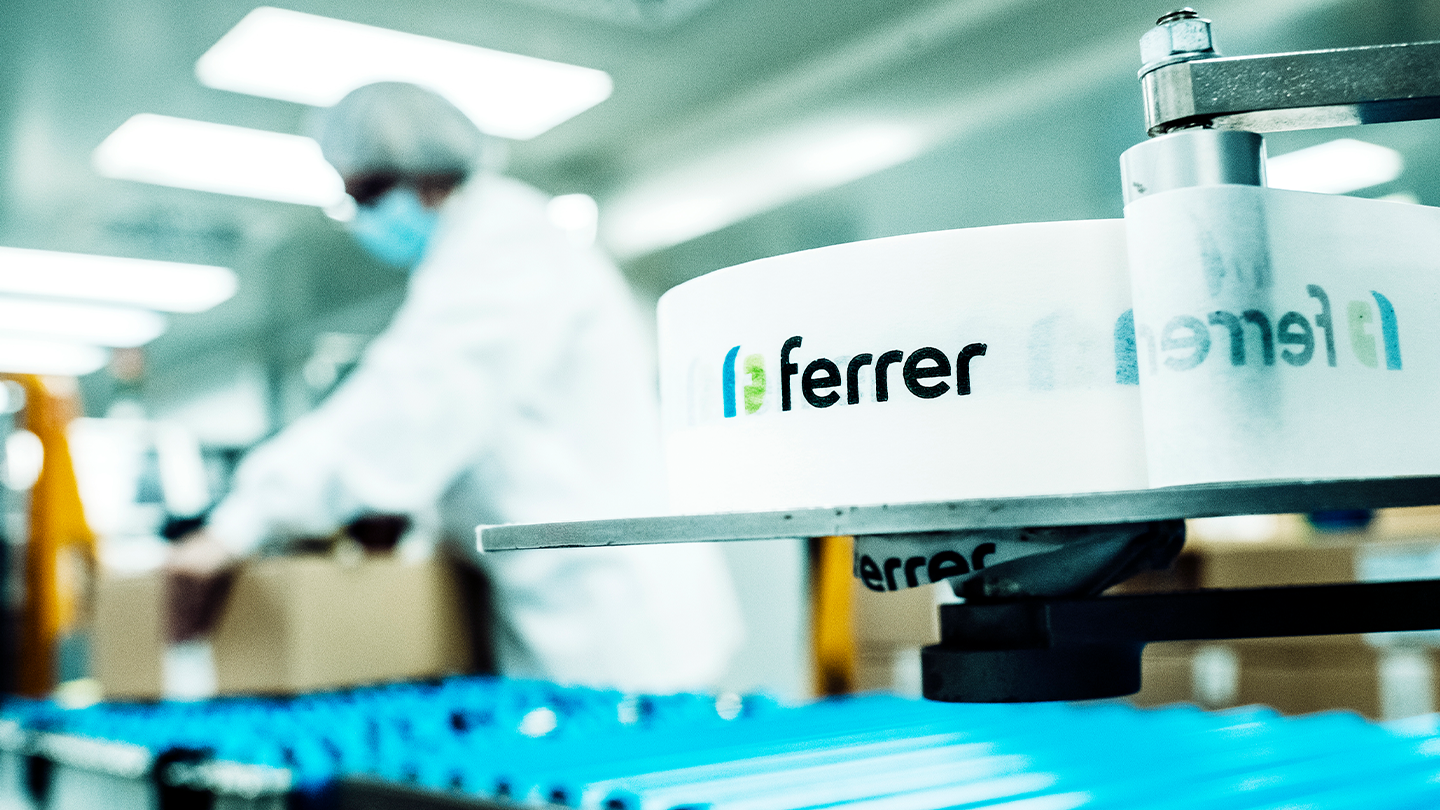 Un rotllo d' etiquetes blanques amb el logotip de Ferrer en primer pla dins d' una planta d' envasament farmacèutic amb un operari desenfocat al fons.