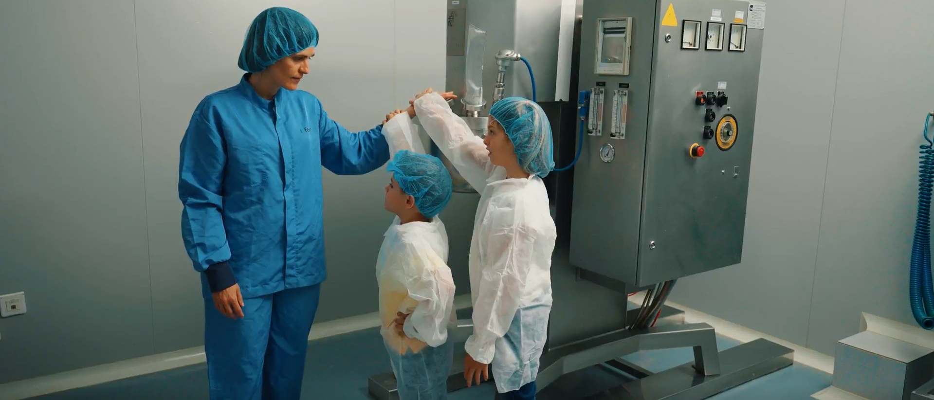 A person from the Ferrer team in a production area, dressed in a uniform and protective cap, showing the space to two children in protective gowns and caps.
