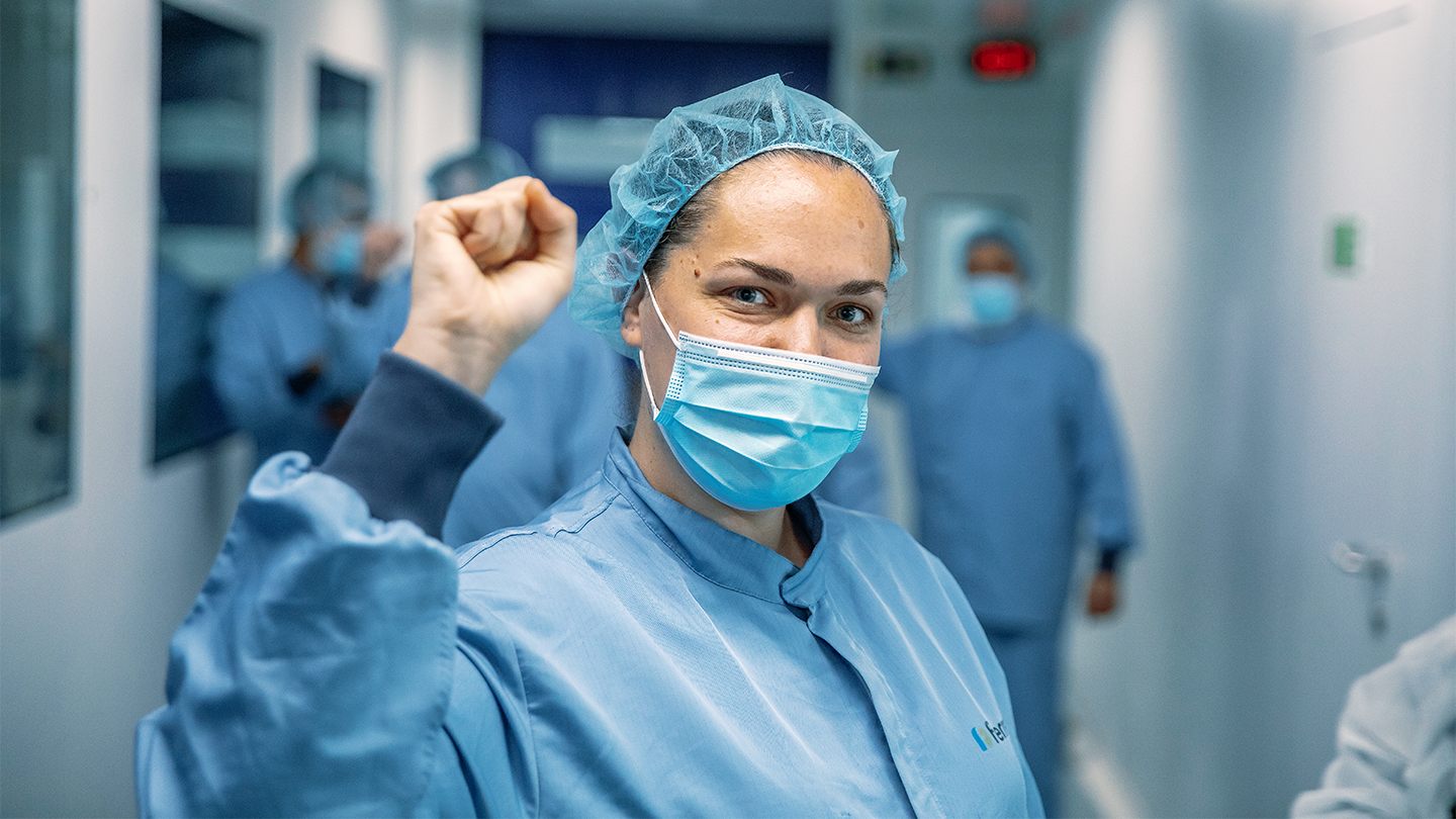 Empleada de Grupo Ferrer con mascarilla y uniforme sanitario celebrando el éxito del equipo en el laboratorio farmacéutico.