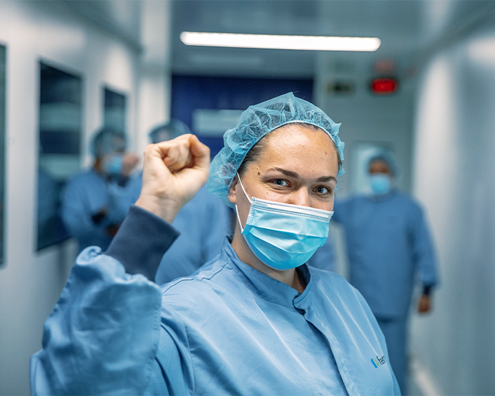 Una profesional de la salud con uniforme, mascarilla y gorro quirúrgico sonríe a la cámara mientras levanta el puño en un pasillo de hospital.