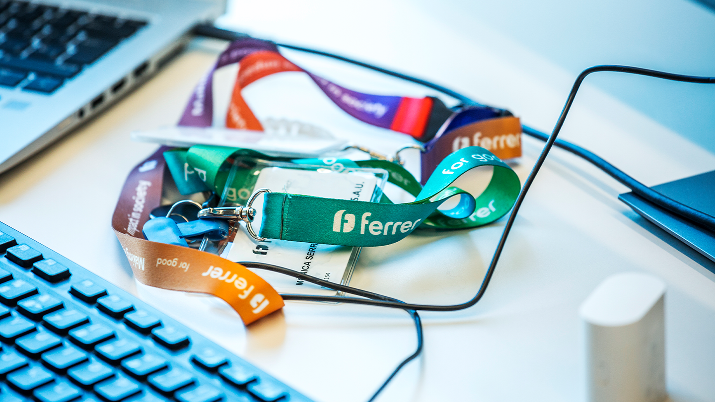 Acreditaciones y lanyards de colores con el logotipo de la empresa Ferrer sobre un escritorio de oficina.
