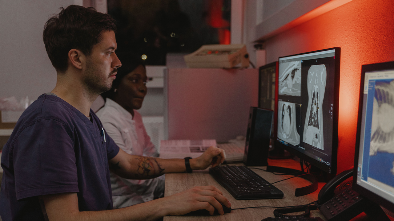 Dos profesionales médicos analizan con atención imágenes de tomografías y radiografías en múltiples monitores de computadora dentro de un laboratorio iluminado con luz tenue y tonos rojizos.