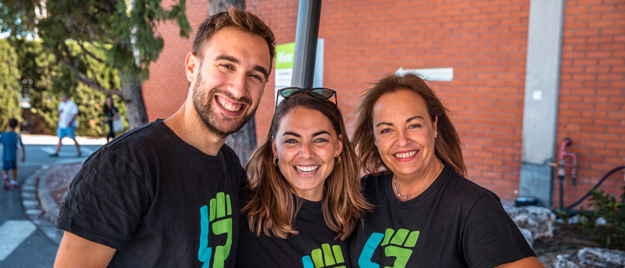 Three volunteers from the pharmaceutical company Ferrer smile and pose together outdoors, wearing black T-shirts with the green logo of a volunteer initiative.