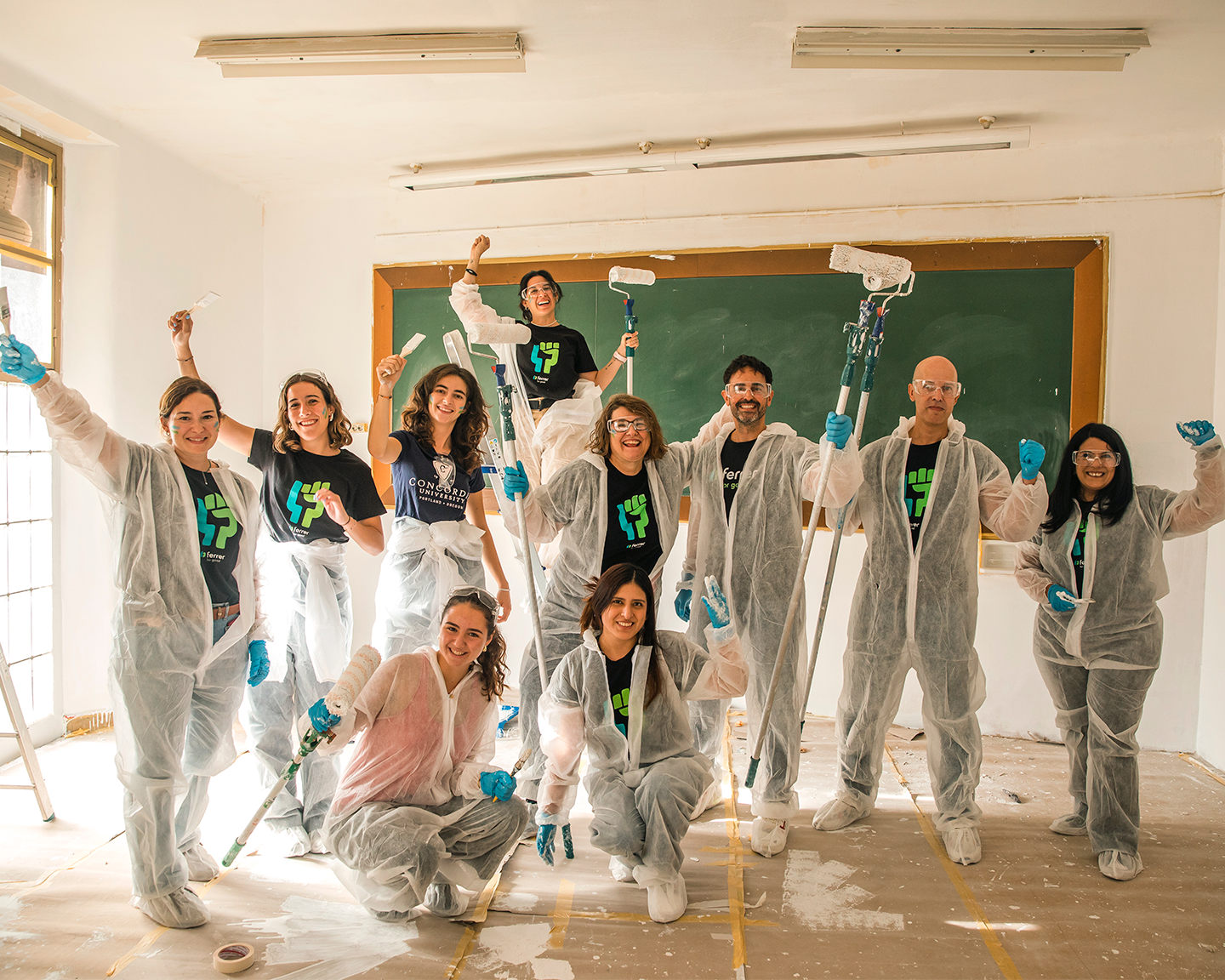 Group of volunteers in a collective action initiative, equipped with protective coveralls and rollers to paint a school classroom with Grupo Ferrer.
