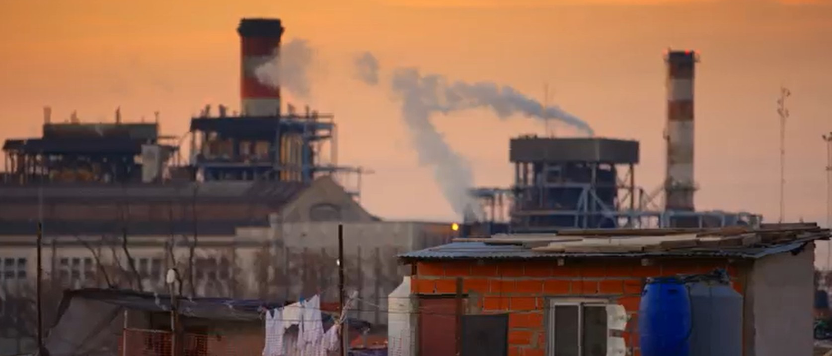 Vista d' una zona industrial al vespre amb fàbriques i xemeneies emetent fum, al costat d' habitatges en primer pla.