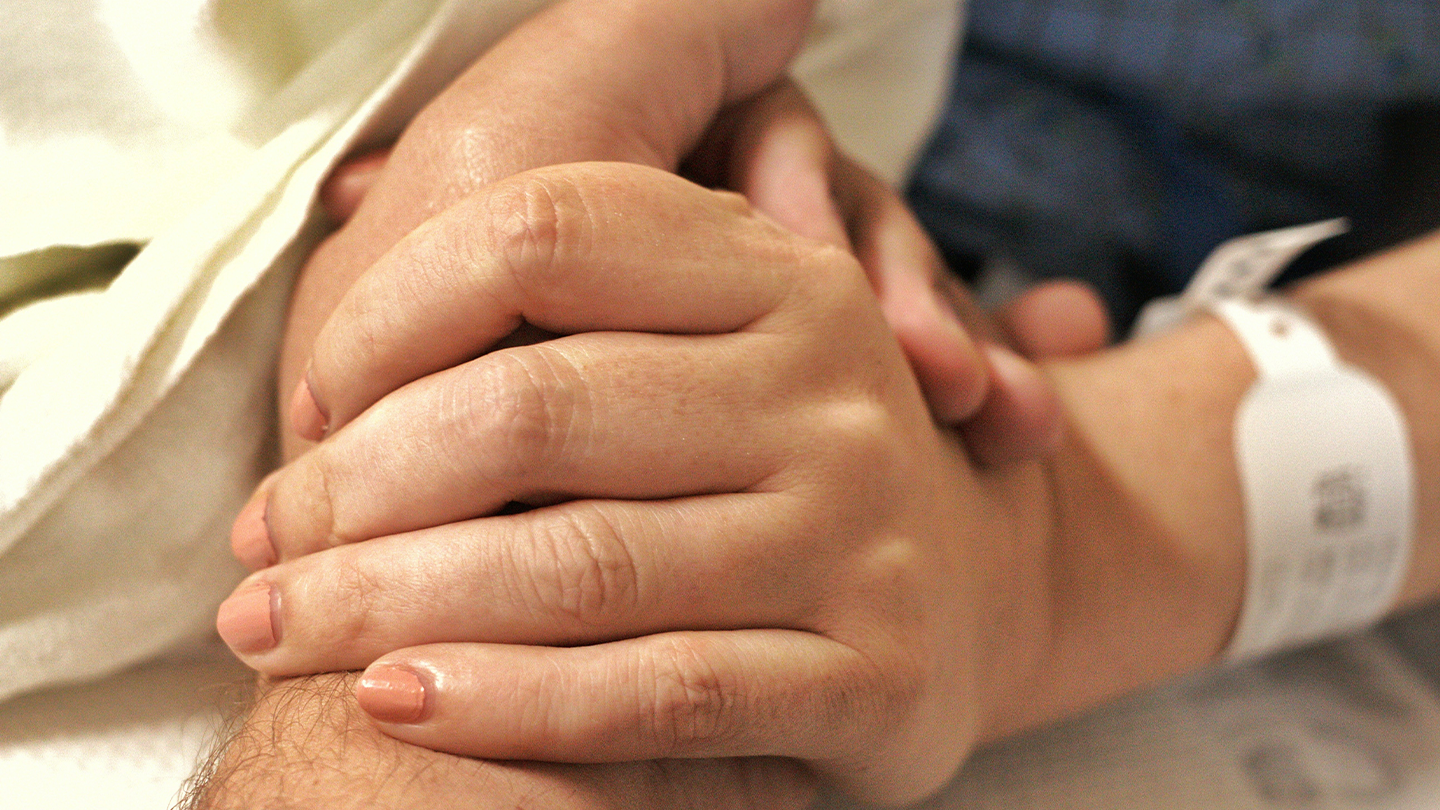 Primer plano de dos personas sosteniendo con afecto la mano de un paciente con un brazalete de identificación en la cama de un hospital.