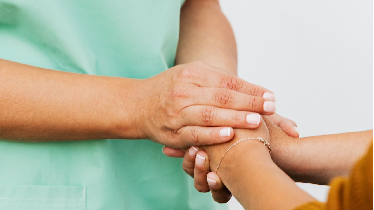 Primer plano de una profesional de la salud en uniforme verde sosteniendo con empatía la mano de un paciente.