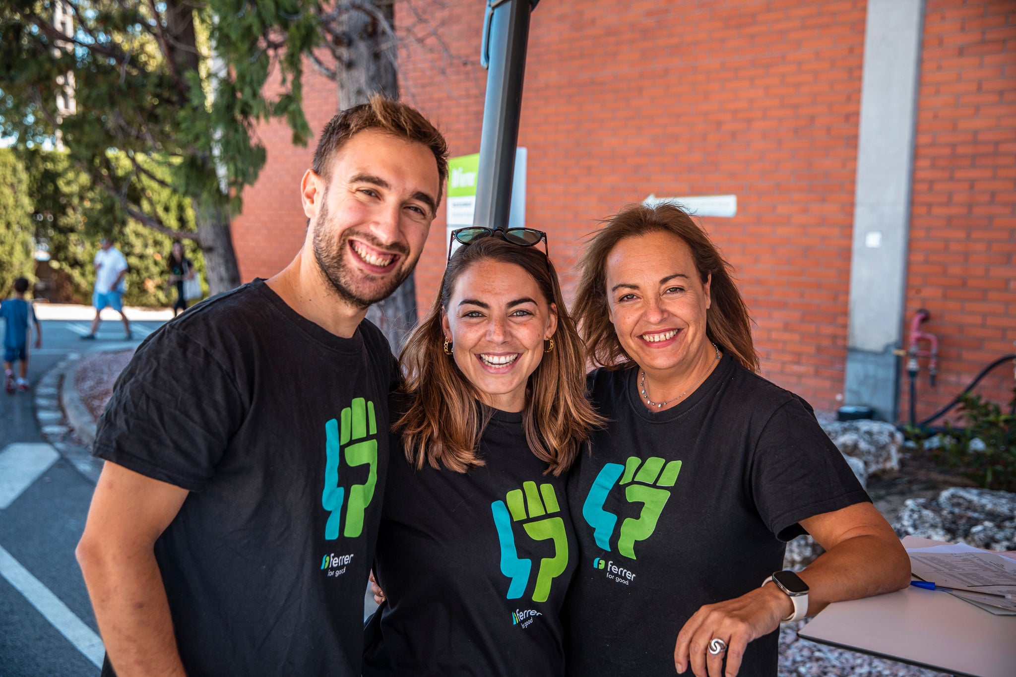 Tres voluntaris de l'empresa farmacèutica Ferrer somriuen i posen junts a l'aire lliure, vestint samarretes negres amb el logo verd d'una iniciativa de voluntariat.