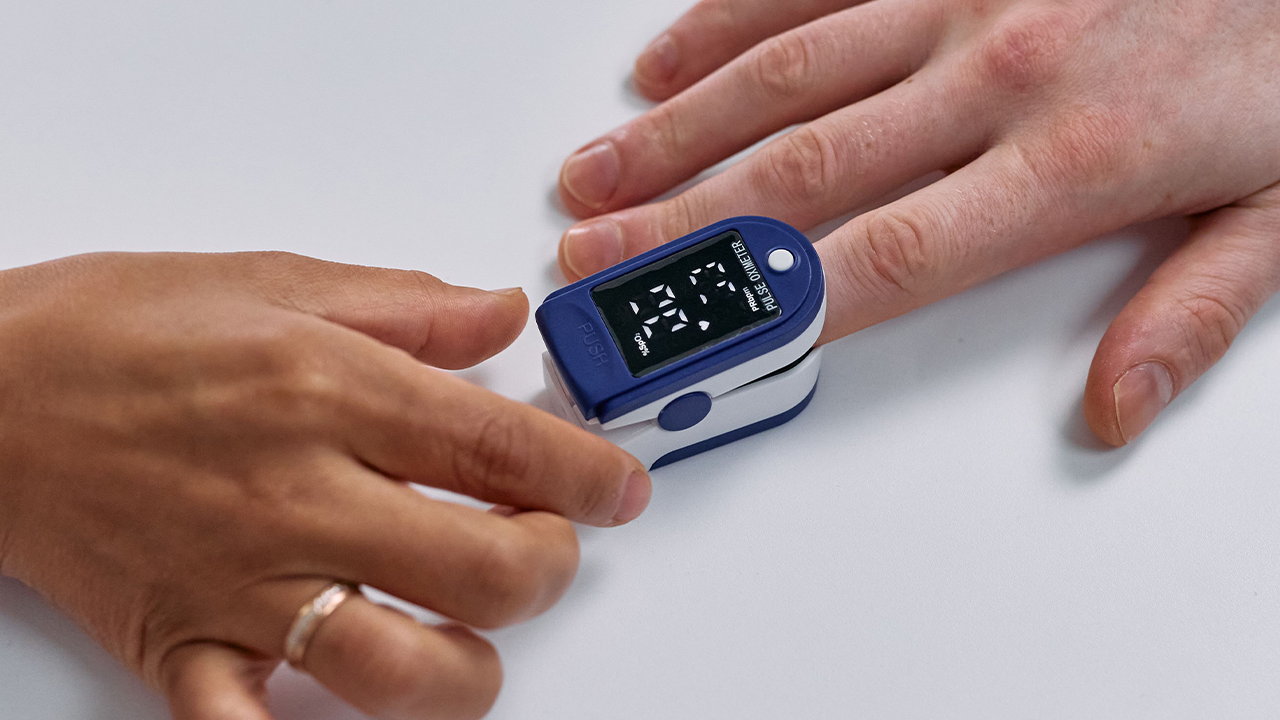 Top view of a person placing a blue and white digital pulse oximeter on another person's index finger on a white surface.