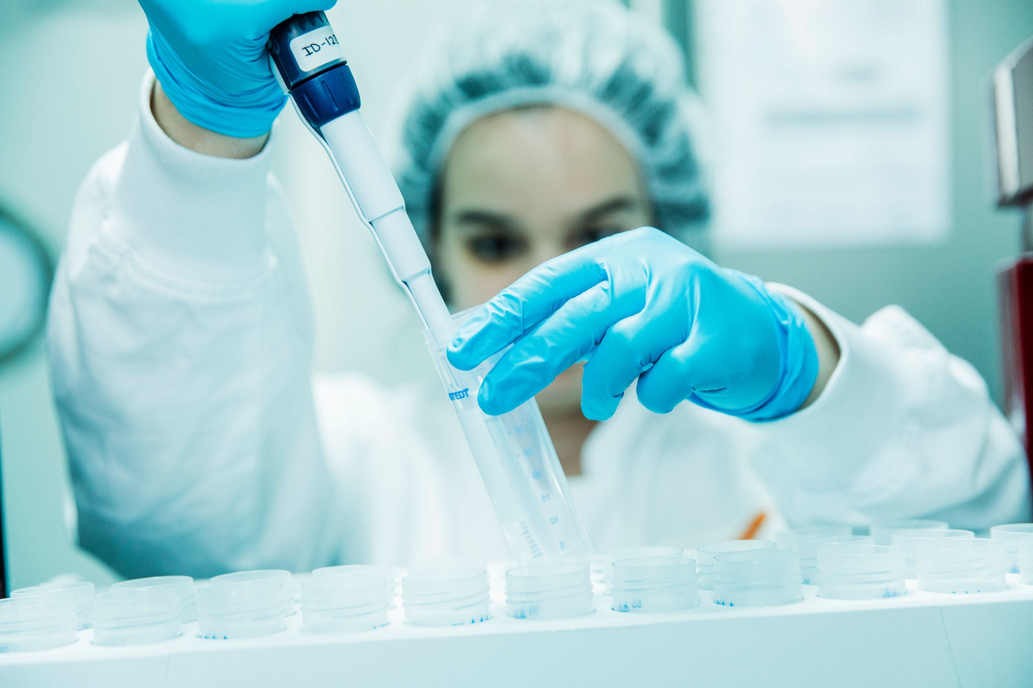 A scientist wearing blue gloves, a gown and a protective cap uses a precision pipette on vials in a laboratory.