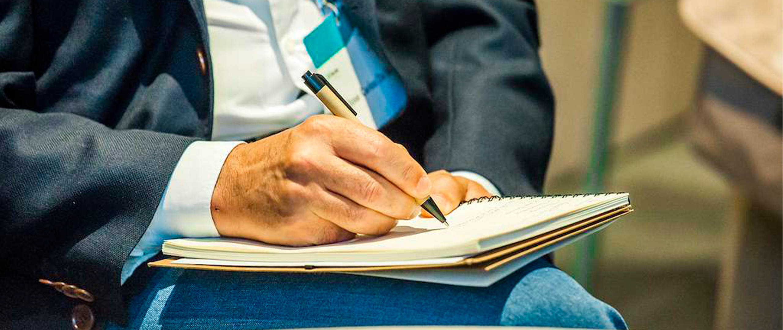 Close-up of a person in a dark jacket taking notes with a pen in a notebook resting on his lap during a corporate event or conference.
