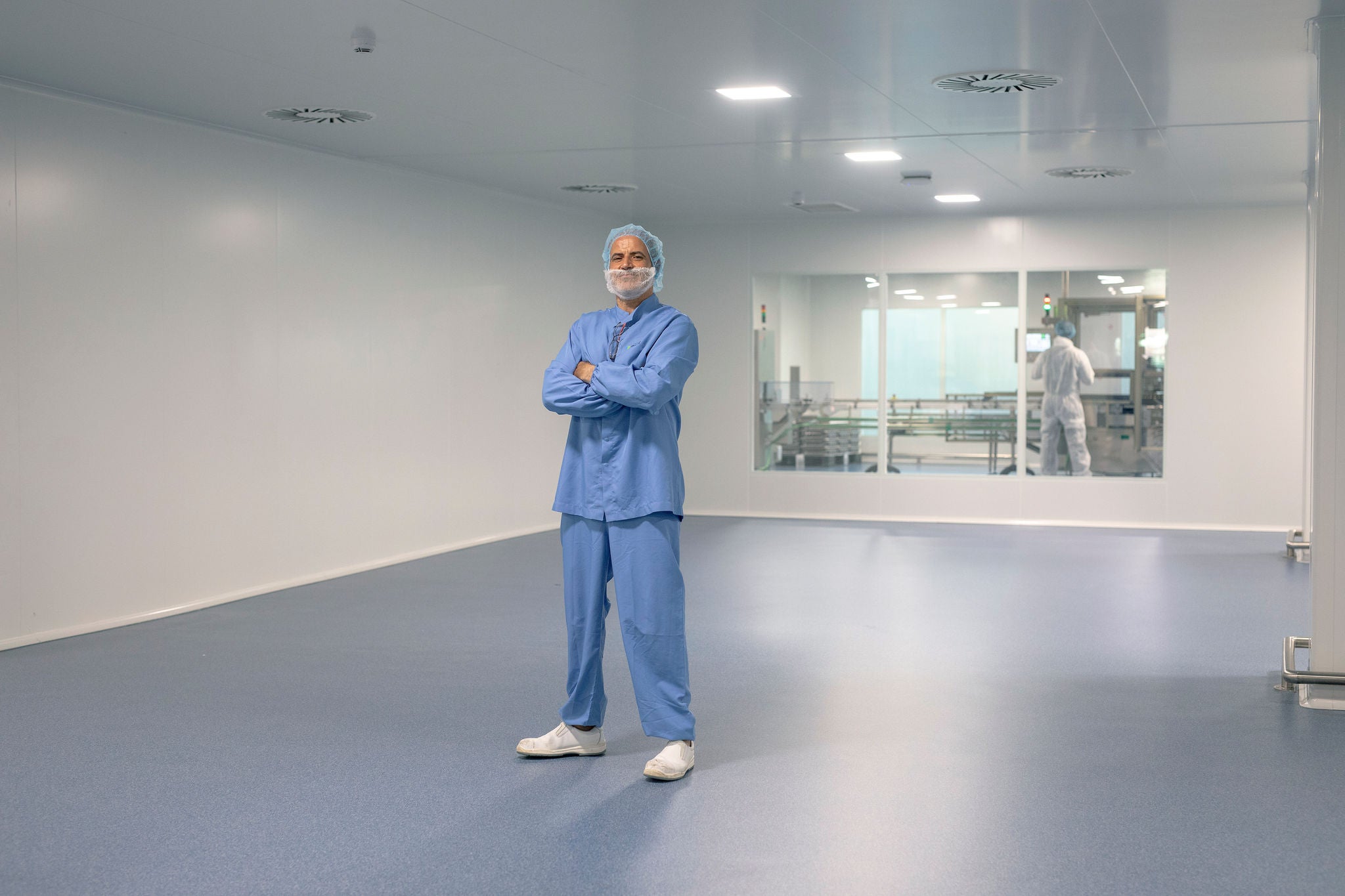 Operari amb uniforme blau de protecció, gorra i mascareta posa amb els braços creuats en una sala blanca industrial d'un laboratori o planta farmacèutica.