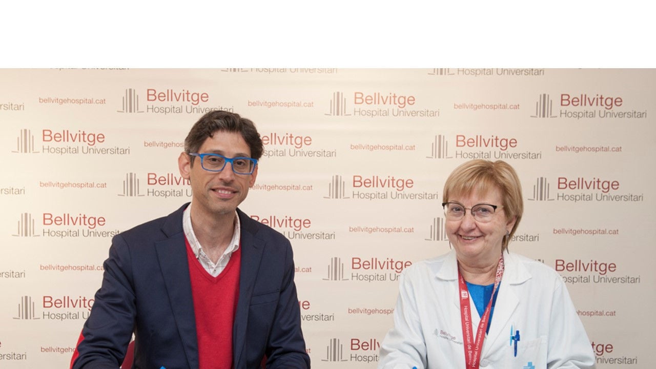  Two health professionals pose smiling in front of a corporate panel at Bellvitge University Hospital.