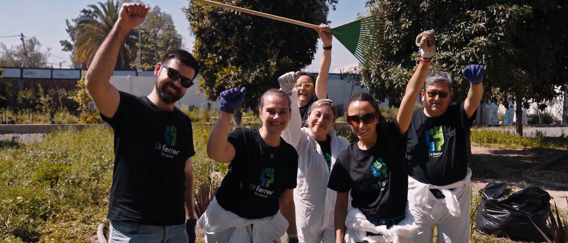 Ferrer volunteers smile and wave to the camera during an outdoor clean-up activity, equipped with gloves and collection bags.