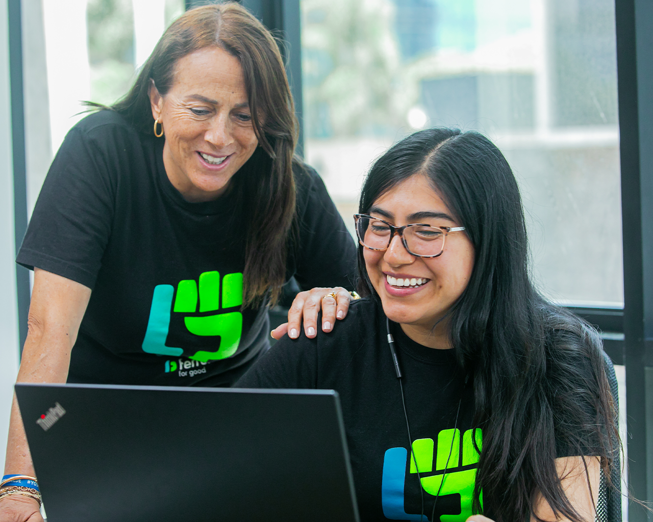 Dos personas del equipo de Ferrer trabajan juntas frente a un portátil, sonriendo en un entorno de oficina y vistiendo camisetas corporativas.