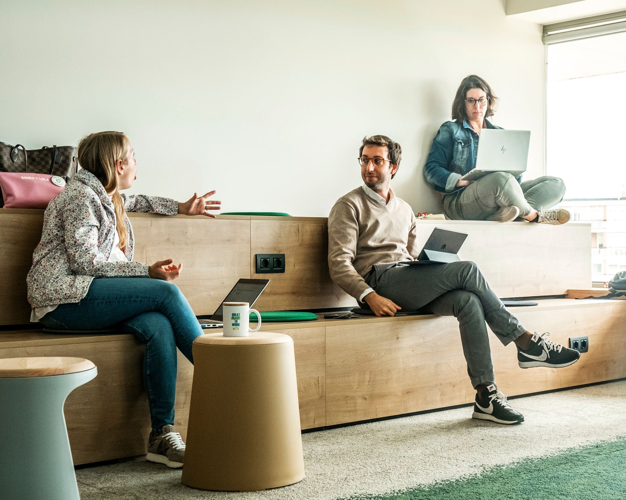 Tres profesionales de Ferrer colaboran en un espacio de trabajo informal, conversando y utilizando portátiles en un entorno de oficina.