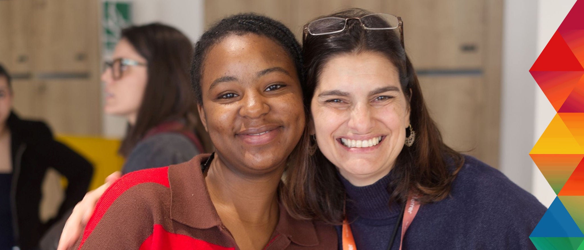 Dos mujeres sonriendo y abrazadas en un entorno interior, con un diseño geométrico de colores en el lateral derecho de la imagen.