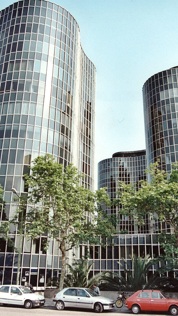 Vista en perspectiva de diverses rascades modernes cilíndricos amb façanes envidrades, situats darrere d' arbres i cotxes estacionats.