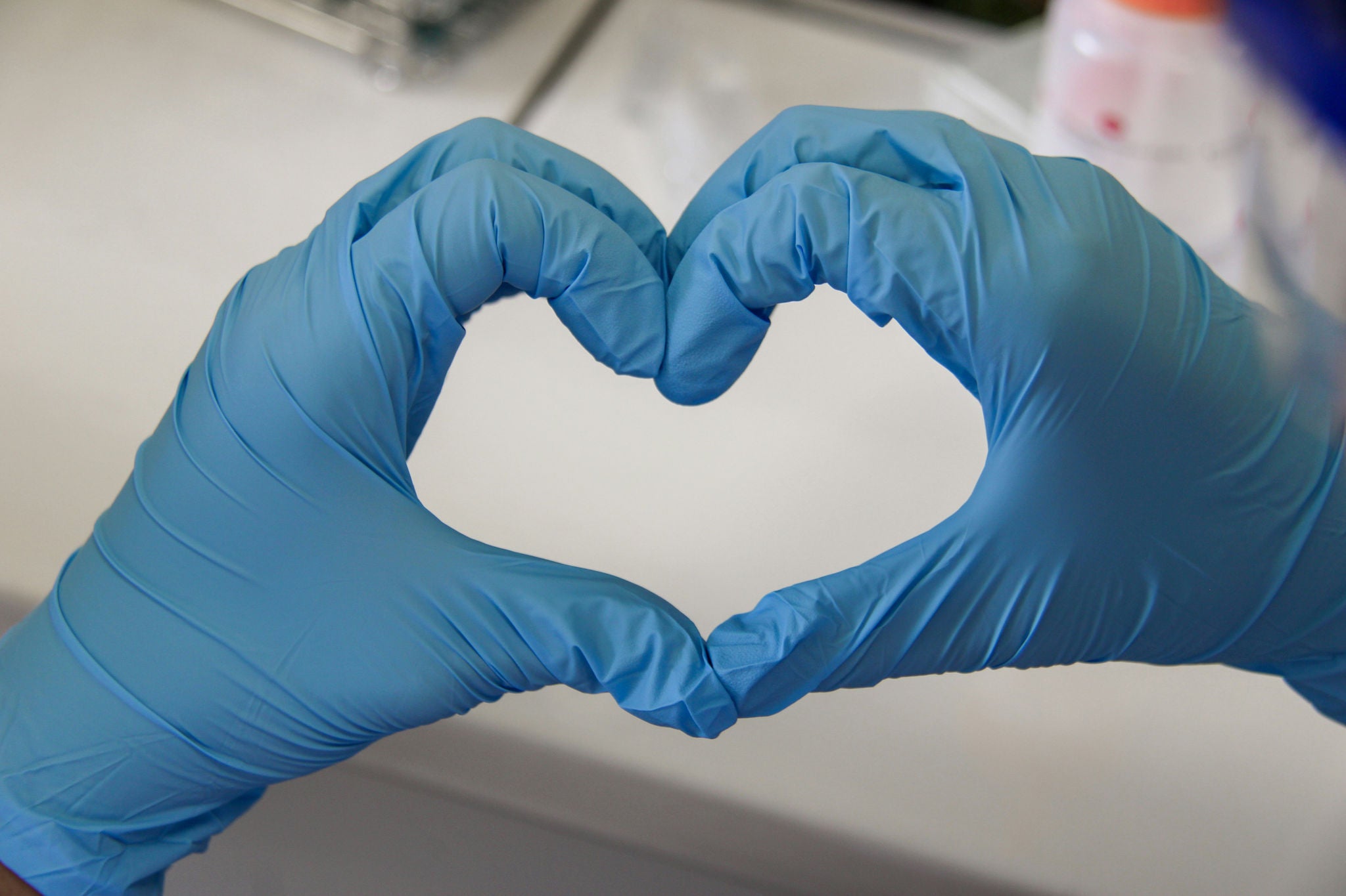 Manos con guantes azules forman un corazón en un entorno de laboratorio, representando el compromiso de Ferrer con el cuidado, la ciencia y la salud.