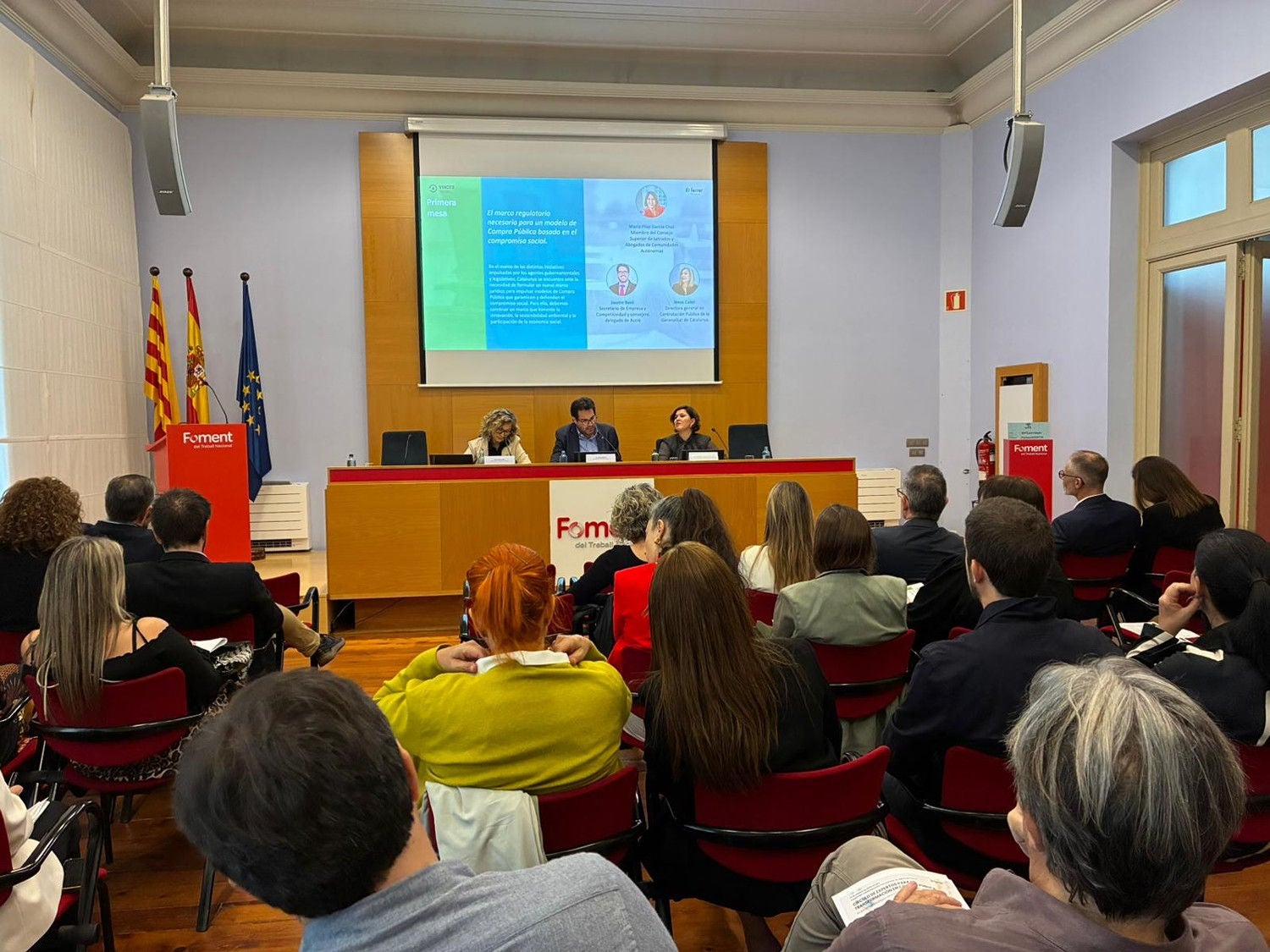 Ponencia en una jornada institucional sobre compra pública, con una moderadora en atril y cinco participantes sentados en una mesa frente al público, bajo una pantalla de presentación.