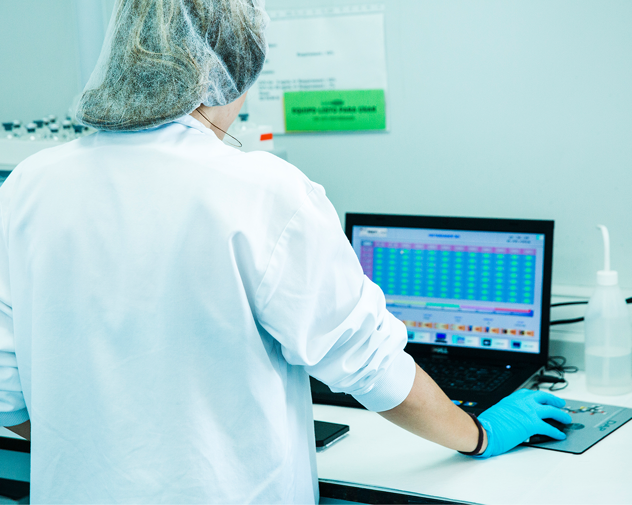 Una técnica de laboratorio con bata blanca y guantes analizando datos en un ordenador en las instalaciones de la empresa Ferrer.