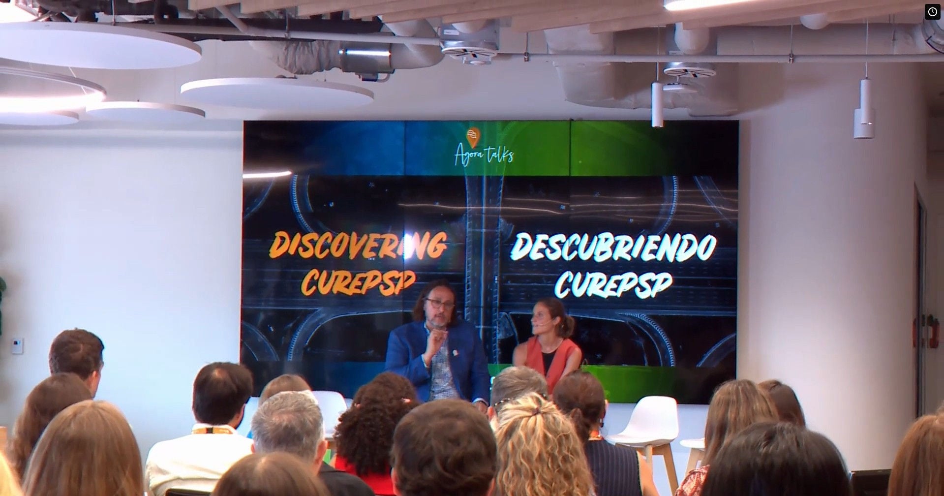 Esdeveniment corporatiu de Ferrer amb dos ponents a l'escenari durant una xerrada, davant una audiència, amb una pantalla de fons que mostra el missatge "Discovering CUREP5P".