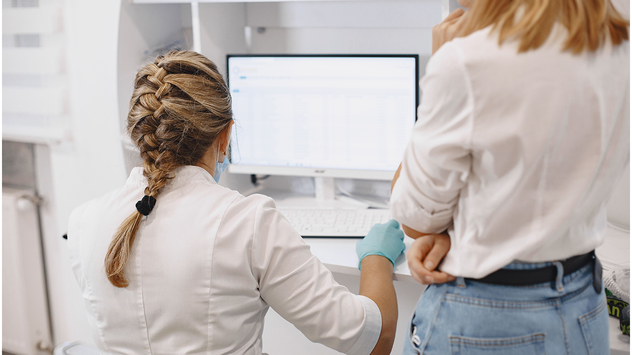 Vista trasera de una profesional médica con el cabello trenzado trabajando frente a una computadora junto a una colega en un consultorio o laboratorio.