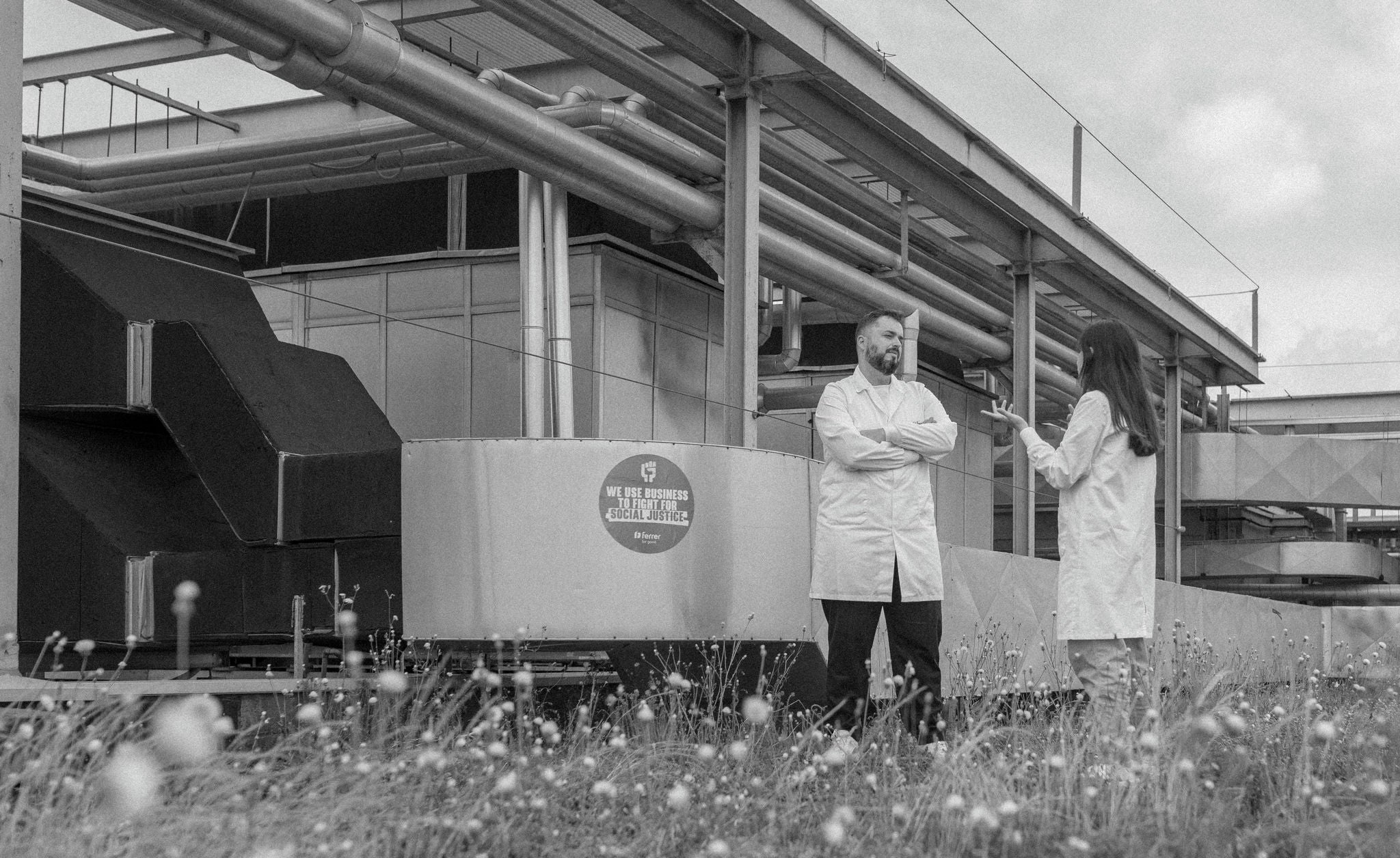 Two professionals in gowns chat in an outdoor industrial area, next to pipes and equipment, with vegetation in the foreground, in a black and white image.